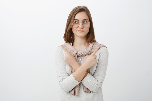 Waist-up shot of relaxed good-looking feminine girl in glasses with short brown hair crossing arms on body pointing in different sides left and right making choice or decision over gray wall