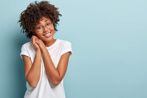 Снимок довольной темнокожей женщины с вьющейся прической, держит обе руки возле лица, имеет нежную улыбку, дружелюбное выражение лица, тронутый комплиментом, носит модели белых футболок на синей стене.