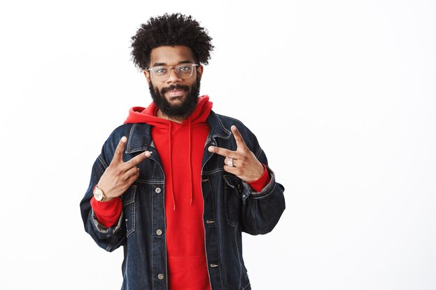 Waist-up shot of satisfied and friendly smiling african american bearded man with afro hairstyle in glasses, denim jacket over red hoodie grinning at camera and showing victory sign over gray wall