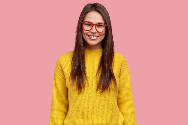 Waist up shot of satisfied pretty brunette young woman with happy expression, wears spectacles and yellow casual jumper, models against pink background