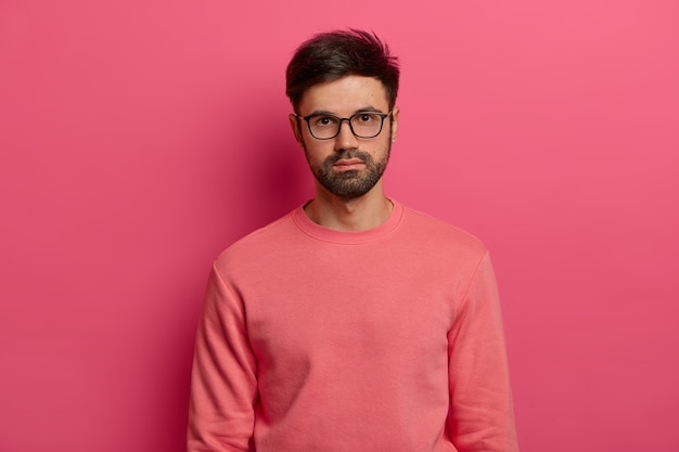 Waist up shot of serious male manager or freelancer looks  with calm expression, focused somewhere, comes on job interview, wears transparent glasses and sweater, poses over rosy wall