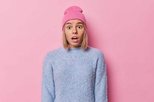 Waist up shot of shocked blonde European woman looks shocked and impressed stands speechless gasps from fear wears casual jumper hat isolated over pink studio background Human reactions concept