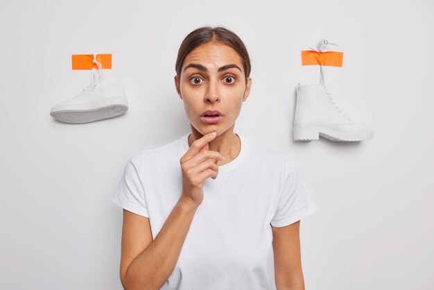 Waist up shot of shocked brunette woman keeps hand on chin stares at camera has stupefied expression dressed in casual basic t shirt poses against white background with plastered sneaker and boot
