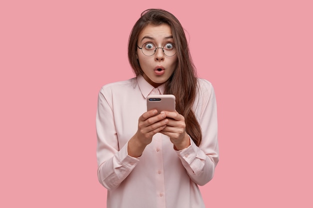Waist up shot of shocked Caucasian lady holds mobile phone, surprised to read spam, made mistake while typing message, recieves high phone bills