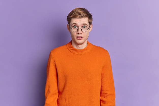 Waist up shot of shocked european man keeps mouth opened holds breath from amazement hears incredible news wears round spectacles and orange jumper