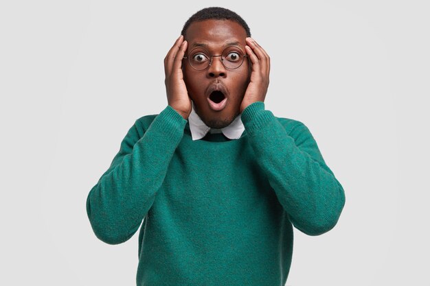 Waist up shot of shocked terrified black masculine adult gasps and stares in stupor, keeps both hands on head, dressed in green jumper and white shirt