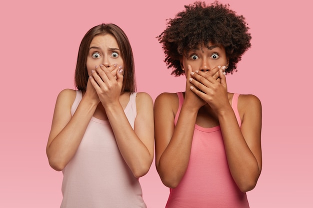 Waist up shot of shocked two women cover mouthes with both hands