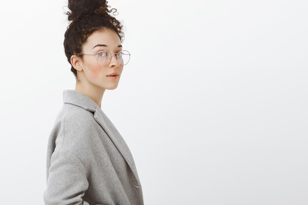 Waist-up shot of smart confident stylish girl with curly hair combed in bun, standing in profile and gazing in glasses