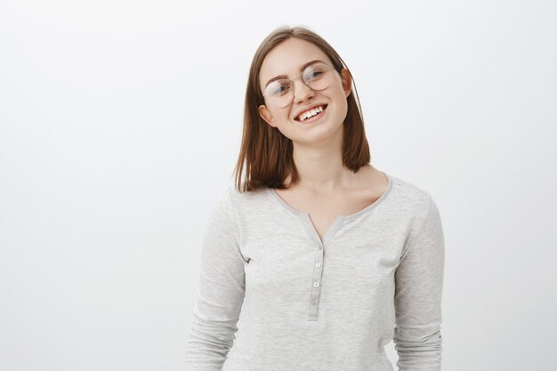 Waist-up shot of smart creative charming young woman with short cute haircut tilting head joyfully smiling broadly wearing glasses and casual white blouse posing happily over grey wall