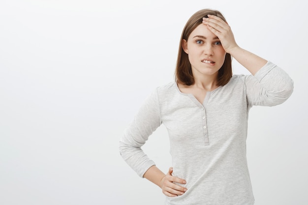 Waist-up shot of smart creative female coworker in white blouse holding hand on forehead biting lower lip being concerned and troubled facing hard situation trying find solution over gray wall