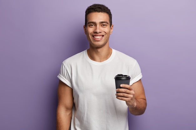 Waist up shot of smiling cheerful handsome guy holds paper takeaway coffee, enjoys spare time with friend, drinks aromatic beverage, wears white mock up t shirt, models against purple wall