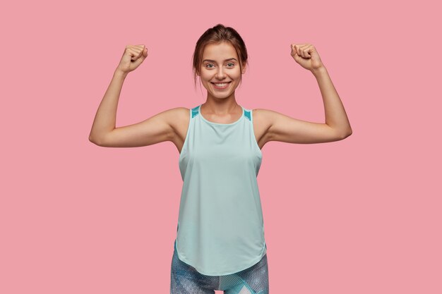 Waist up shot of sporty woman raises hand to show her muscles