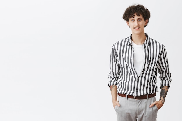 Free photo waist-up shot of stylish modern young hipster guy in striped shirt and gray pants holding hands in pockets with moustache dark curly hair and tattoos dressing up for date over gray wall