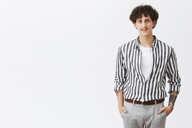 Waist-up shot of stylish modern young hipster guy in striped shirt and gray pants holding hands in pockets with moustache dark curly hair and tattoos dressing up for date over gray wall