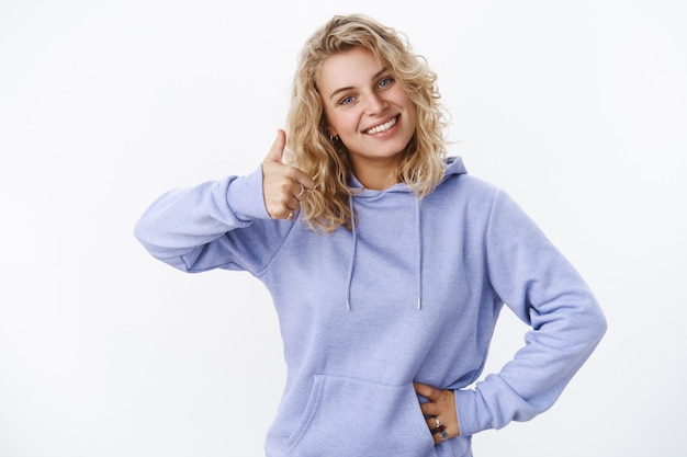 Waist-up shot of supportive pleasant and satisfied attractive european fair-haired female in hoodie tilting head joyfully showing thumb up in like and approval, recommend cool product over white wall