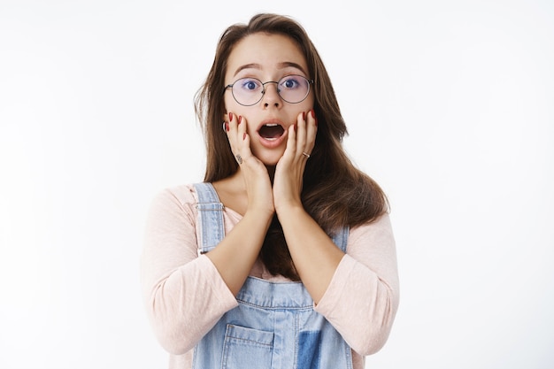 Free photo waist-up shot of surprised and amazed young cute female geek in glasses dropping jaw saying wow holding hands on cheeks impressed and astonished with great awesome news over gray wall. copy space