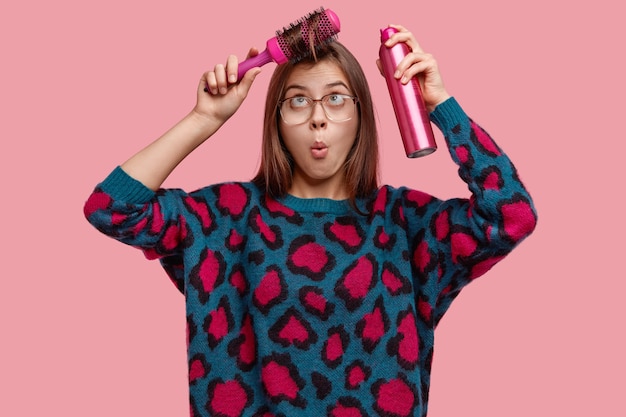 Waist up shot of surprised awkward woman combes fringe, sprays for making haircut, wears big spectacles, sweater