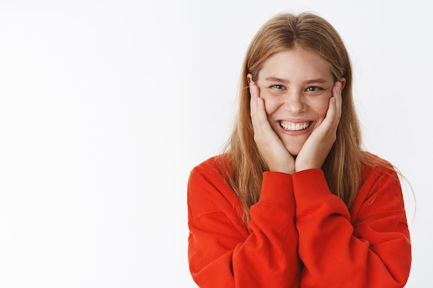 Waist-up shot of surprised happy charming young female with freckles and beautiful blue eyes pressing hands to cheeks from amazement and joy