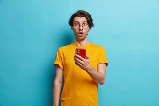 Waist up shot of surprised hipster guy reacts emotionally on shocking news holds mobile phone