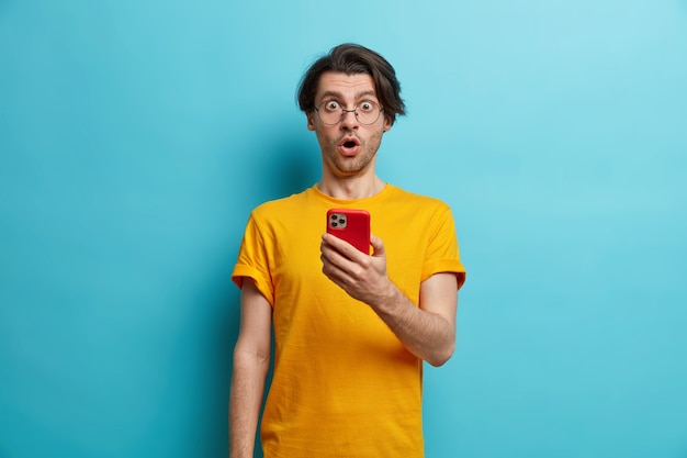 Waist up shot of surprised hipster guy reacts emotionally on shocking news holds mobile phone