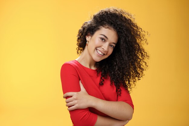 Waist-up shot of tender feminine and gentle woman with curly hairstyle combed to right side, tilting head and smiling flirty making romantic gazed at camera hugging herself over yellow background.