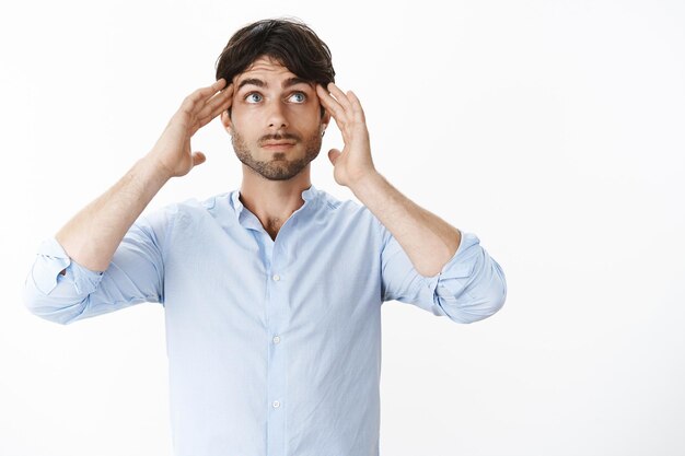 Waist-up shot of thoughtful good-looking male employee in shirt holding hands on temples as looking at upper right corner trying invent idea or plan focusing on project over gray wall.