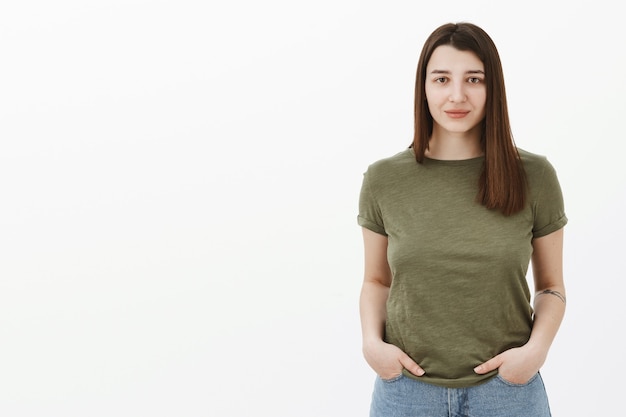 Waist-up shot of trustworthy calm and cute friendly-looking young 20s woman holding hands in pockets relaxed and casual pose smiling cute with slight grin over gray wall