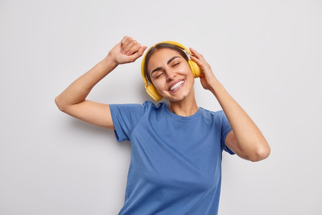 Waist up shot of upbeat cheerful young European woman moves with rhythm of music wears wireless headphones and casual blue t shirt smiles toothily dances upbeat isolated over white background