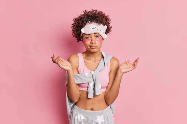 Waist up shot of woman spreads palms has hesitant expression applies collagen patches under eyes to reduce puffiness dressed in casual pajama isolated over pink studio wall