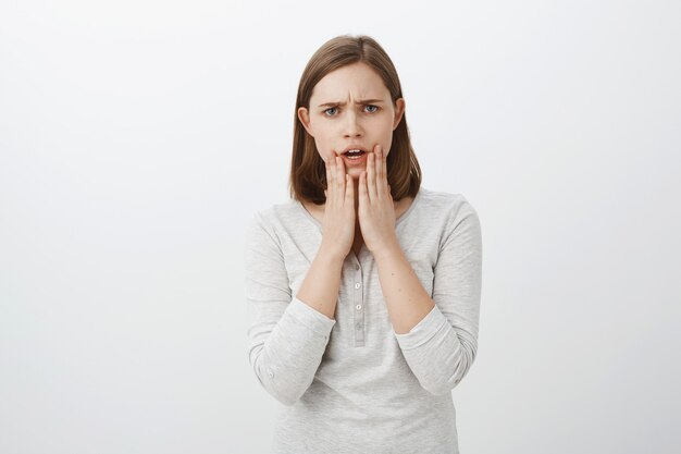 Waist-up shot of worried nervous young brunette woman frowning looking concerned holding palms on chin expressing empathy while hearing shocking event happened to friend over gray wall