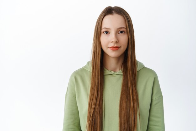 Waist up shot of young beautiful woman with long healthy hair, natural nude makeup, smiling and looking determined at front, standing in hoodie against white wall