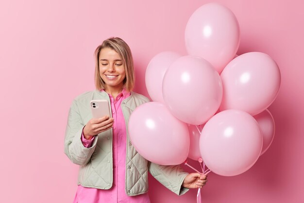 Waist up shot of young European smiles toothy uses mobile phone gets message of congratulations dressed in jacket holds bunch of inflated balloons isolated over pink background. Birthday concept