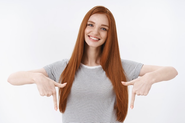 Vita alta tenera bella rossa giovane studentessa inclinando la testa sorridendo felici denti bianchi che puntano gli indici soddisfatti verso il basso discutendo con gioia di un prodotto interessante verso il basso