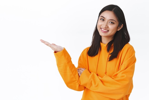 Waistup portrait cute beautiful asian woman in orange hoodie introduce produce banner pointing at blank white space holding product and smiling recommend advertisement white background