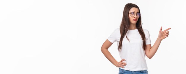 Free photo waistup portrait of uneasy sad and gloomy young brunette woman in glasses looking and pointing finge