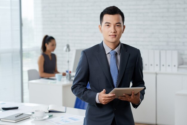 Waistup shot of Asian business man standing in the moddle of the office and holding a digital tablet