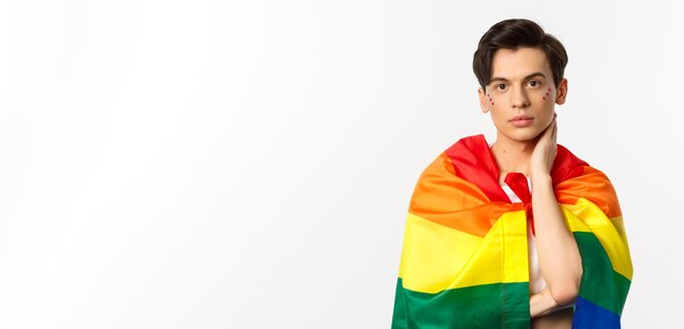 Waistup shot of beautiful gay man with glitter on face wrap himself with rainbow lgbt flag and touch