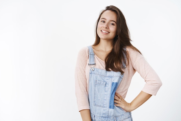 Waistup shot of charming tender and confident female with brown hair in dungarees holding hand on waist and smiling broaldy at camera with assertive selfassured expression over gray background