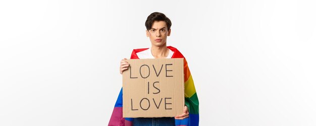 Waistup shot of young lgbtq male activist wearing rainbow flag and holding love is love card sign fo