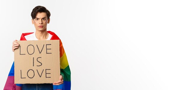 Waistup shot of young lgbtq male activist wearing rainbow flag and holding love is love card sign for pride parade fighting for human rights white background