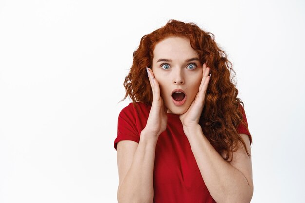 Wait what Shocked redhead girl drop jaw from gossips gasping and staring with disbelief while holding hands on face standing impressed against white background