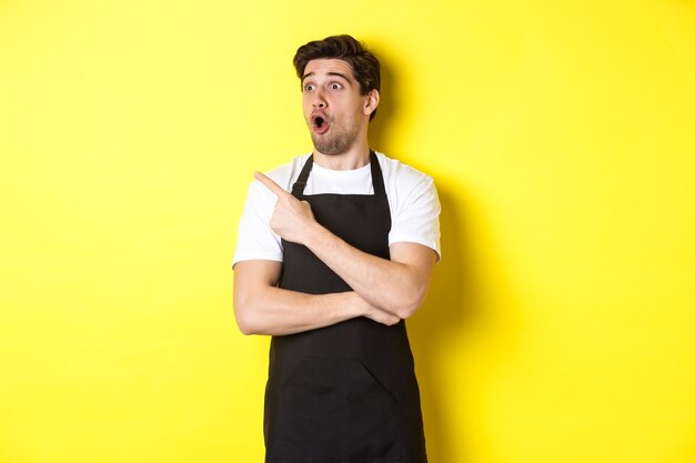 Waiter in black apron checking out promo offer, pointing finger and looking left at your logo, standing over yellow background