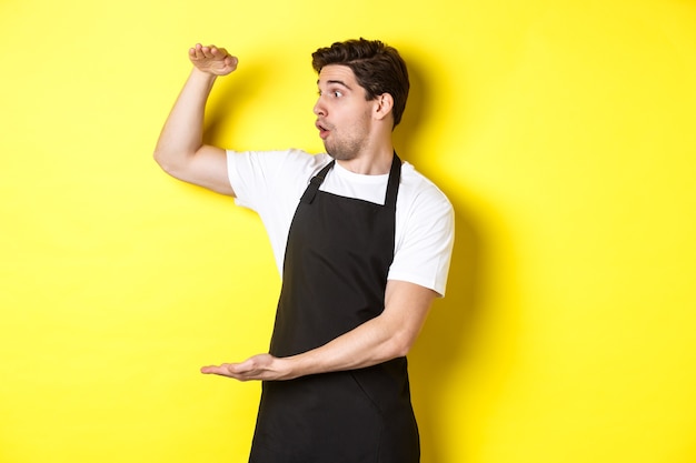 Waiter in black apron looking amazed at something large, holding big object, standing over yellow background.