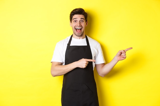 Waiter in black apron pointing fingers right, showing advertisement and smiling excited, standing against yellow background