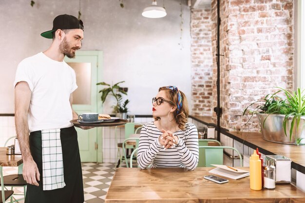 カフェのテーブルに座っている眼鏡のきれいな女性にハンバーガーとコーヒーを与える黒い帽子のウェイター