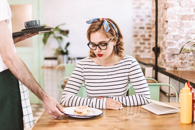 Free photo waiter in black cap giving order to nice lady in eyeglasses which sitting at the table and thoughtfully looking on burger in cafe