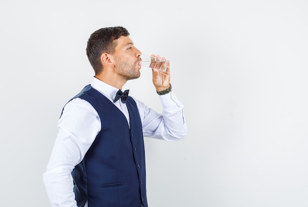 Официант пьет стакан воды в рубашке, жилете и хочет пить.