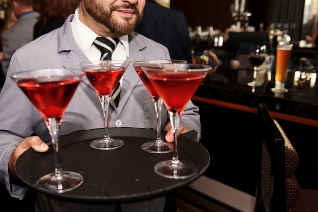 Официант в серой куртке несет коктейльные бокалы с красными напитками