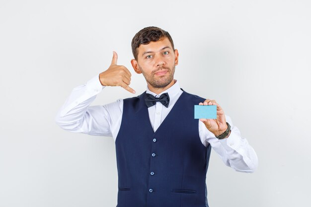 Waiter holding business card with phone gesture in shirt, vest and looking helpful , front view.