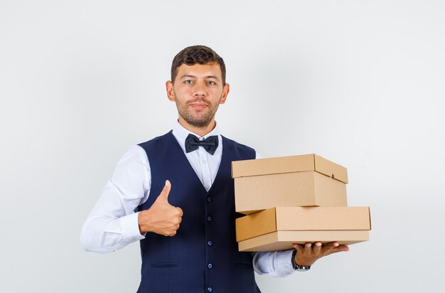 Waiter holding cardboard boxes with thumb up in shirt, vest and looking pleased. front view.